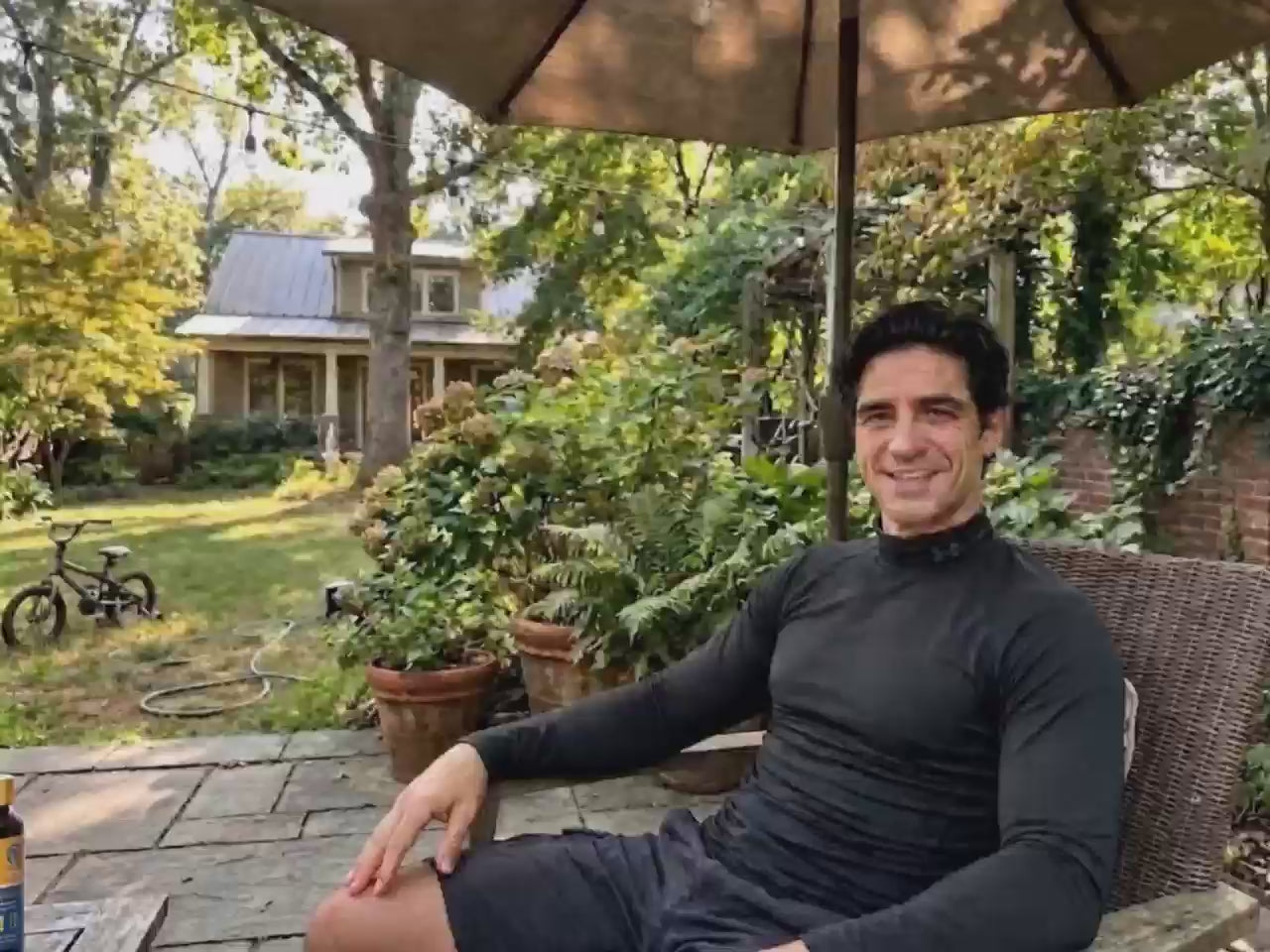 A man relaxing in a sunny backyard under an umbrella, smiling while holding up a BioHarmony Oyster Peptide & Maca bottle. (影片 / video)